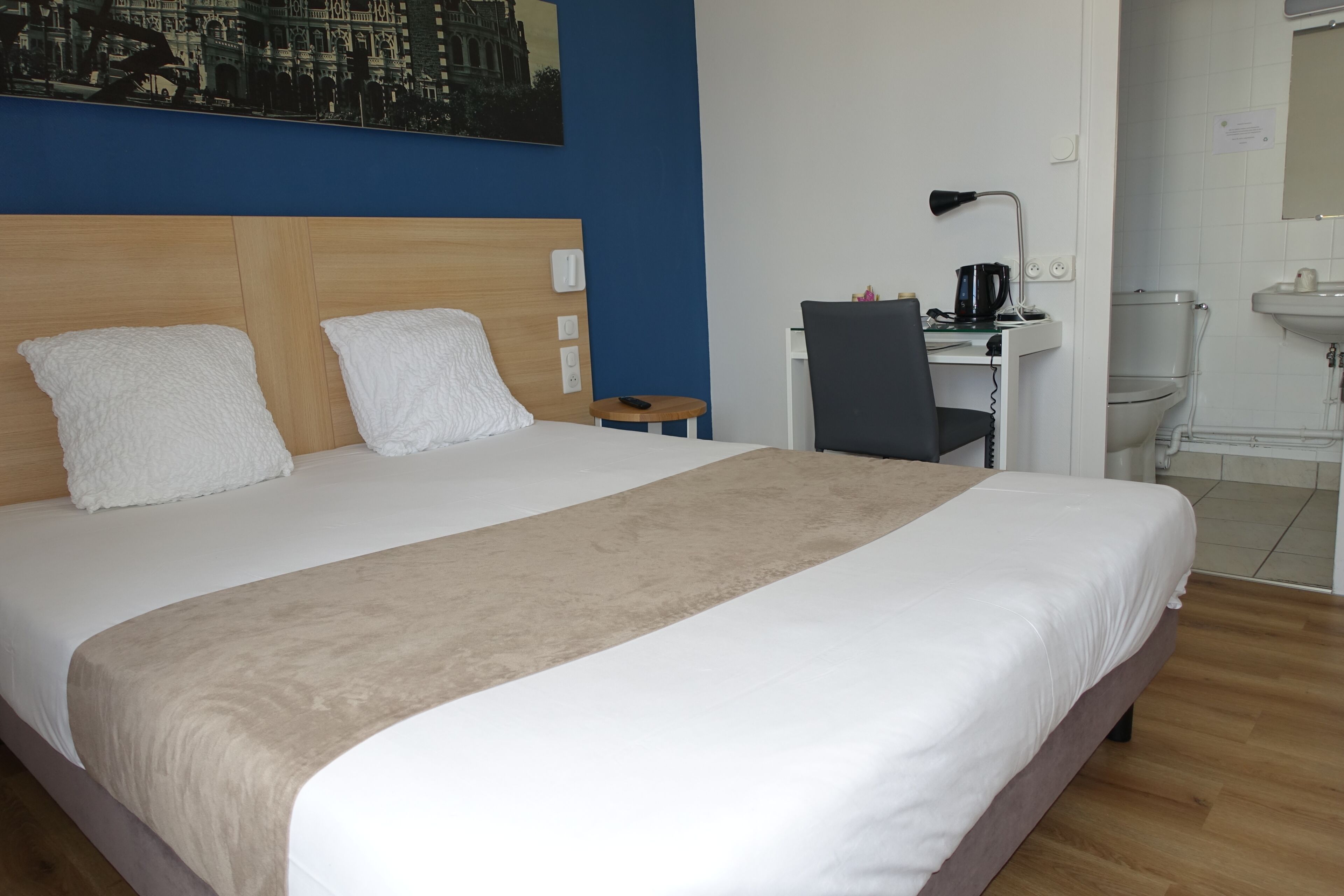 A hotel room with a large bed, a desk with a chair, a small table, a lamp, and a bathroom with a toilet and sink.