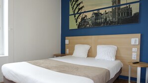 A neatly made bed with a wooden headboard, a bedside table with a remote control, and a wall-mounted light switch. - Hôtel de la Gare (La Roche-sur-Yon)