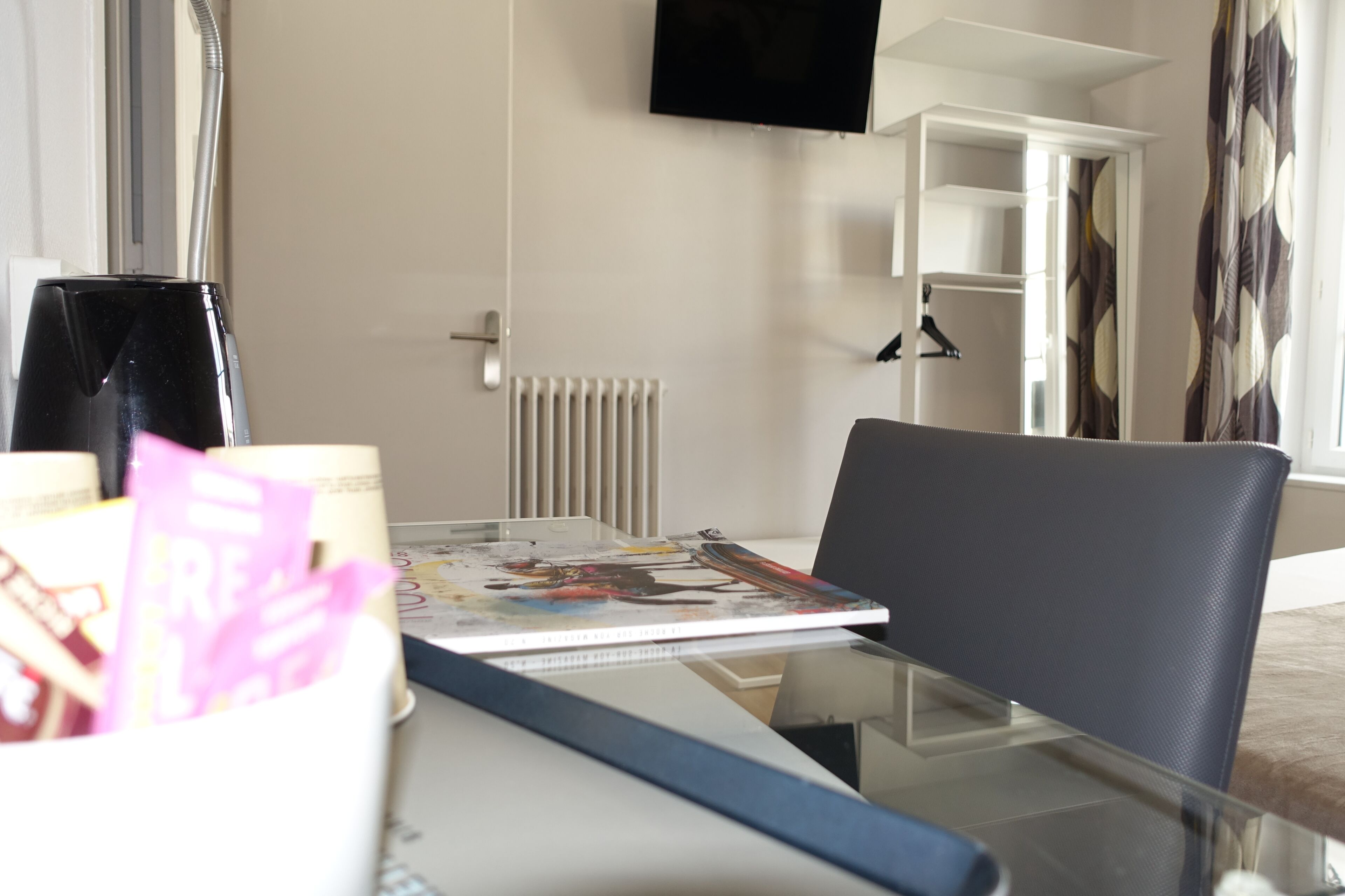 Une chambre d’hôtel avec une table en verre, une chaise noire, un magazine, une machine à café et un téléviseur fixé au mur.