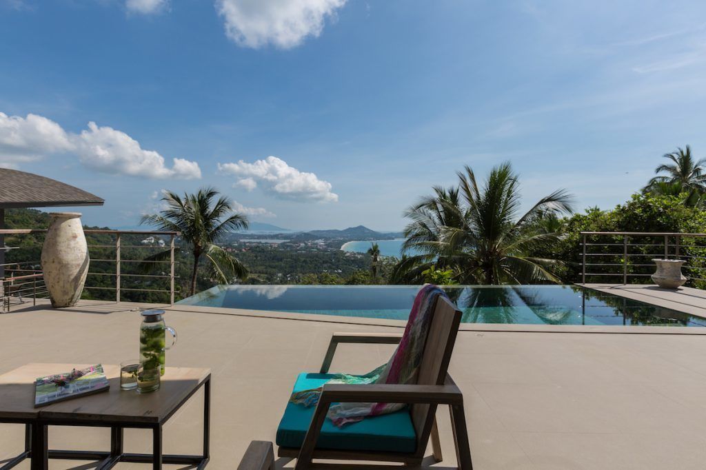 Un espace aménagé au bord de la piscine, avec un fauteuil de relaxation, une table sur laquelle sont posés un magazine et une boisson, et une vue sur un paysage comprenant des montagnes et des palmiers.