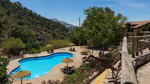 A swimming pool with lounge chairs and umbrellas, surrounded by a stone deck and lush greenery, with a mountainous backdrop. - Calerilla Hotel (La Iruela)