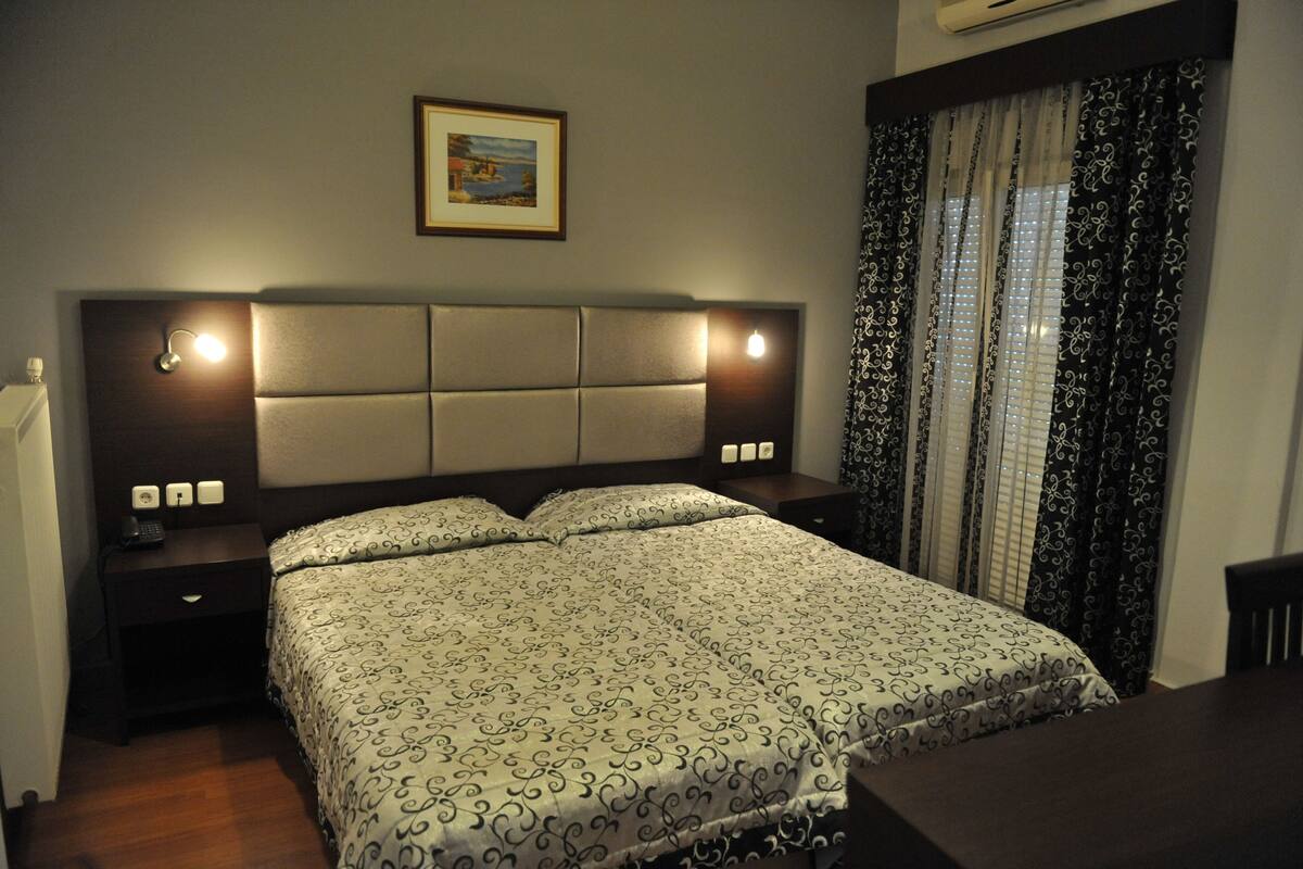 Ein ordentlich eingerichtetes Hotelzimmer mit einem großen Bett, Nachttischen mit Lampen, einem gerahmten Bild an der Wand und einem Fenster mit gemusterten Vorhängen.