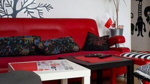 A modern living room with a red sofa, a white coffee table, and a black chair. There is a plant on a metal stand and a wall decoration of a tree. - Art Hostel (Kraków)