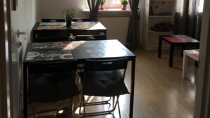 A room with a wooden floor, a dining table with chairs, a bookshelf, and a window with curtains. - Art Hostel (Kraków)