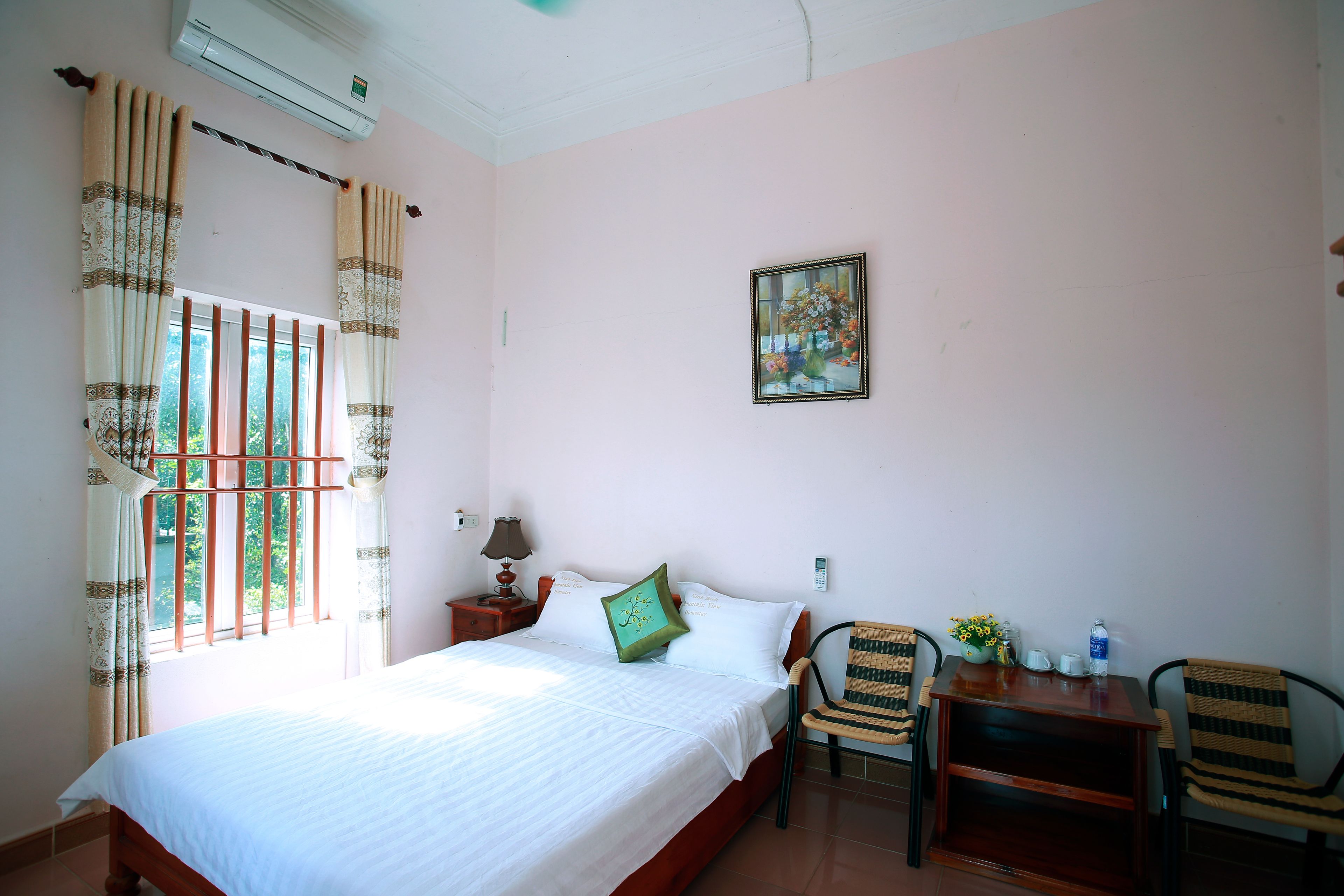 A bedroom with a bed, a chair, a small table, a window with curtains, a wall-mounted air conditioner, and a painting.