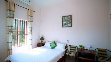 A bedroom with a bed, a chair, a small table, a window with curtains, a wall-mounted air conditioner, and a painting.