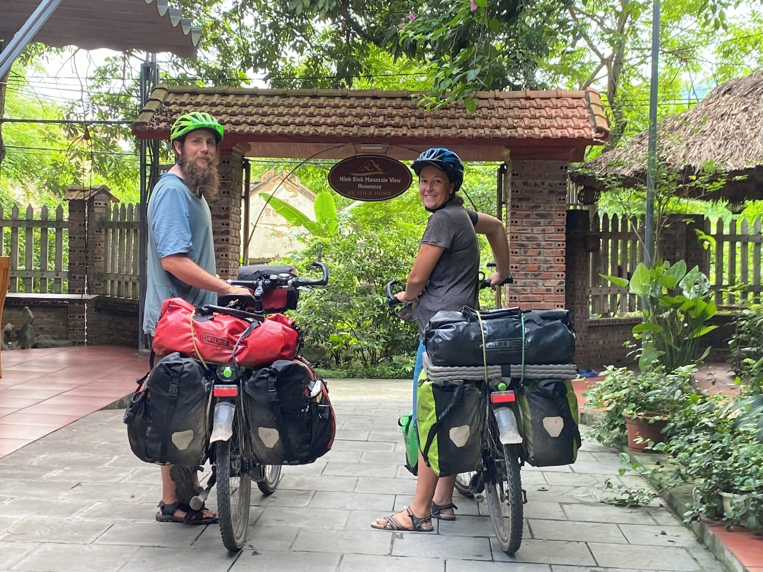 Two people on bicycles with luggage, a brick structure, and a sign indicating a view.