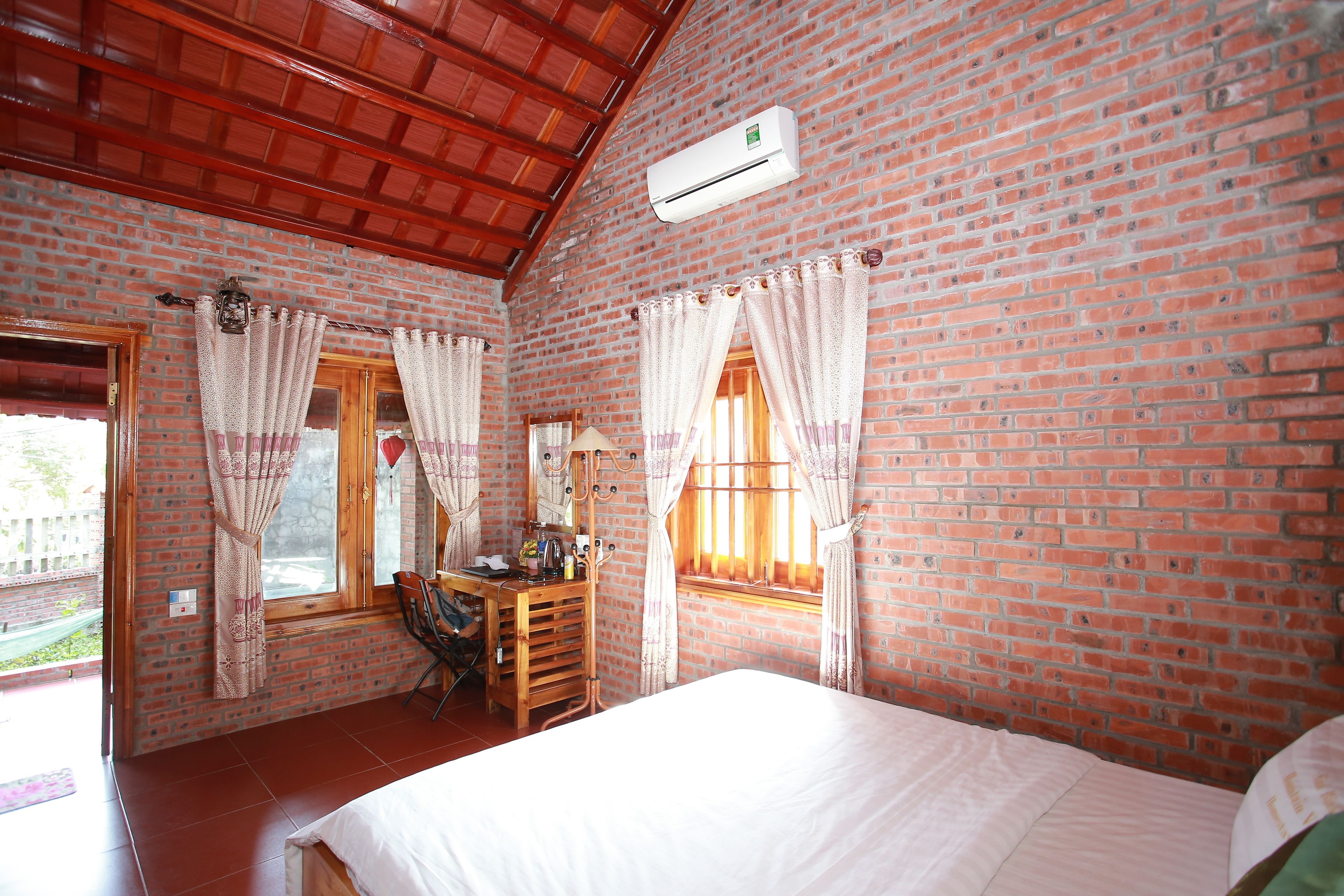 A room with brick walls, a wooden ceiling, a bed, a desk with a chair, and two windows with curtains.