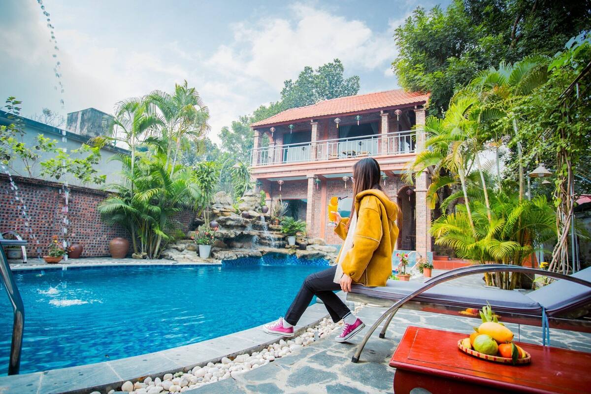A pool area with a person sitting on a bench, a table with fruit, and a building with a balcony in the background.