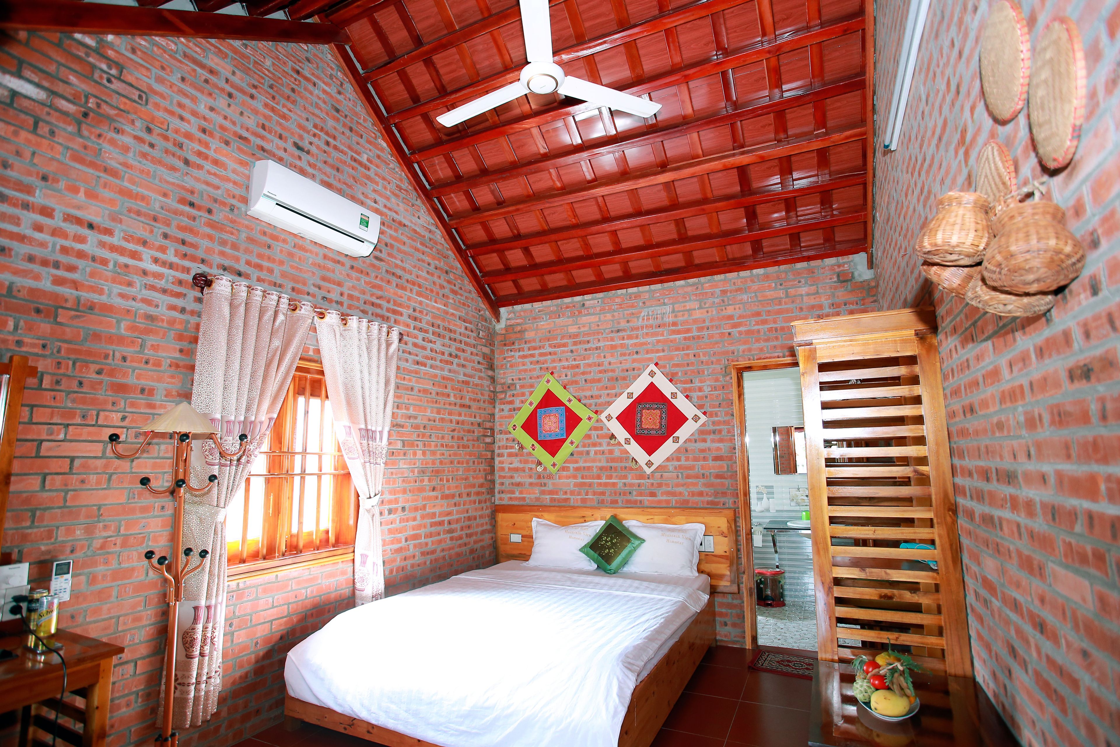 Una habitación con pared de ladrillo, una cama con ropa blanca, un ventilador de techo y una ventana con cortinas.