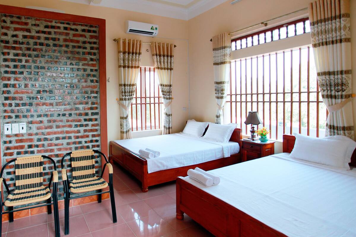 A hotel room with two beds, a brick wall, a window with bars, and a wicker chair.