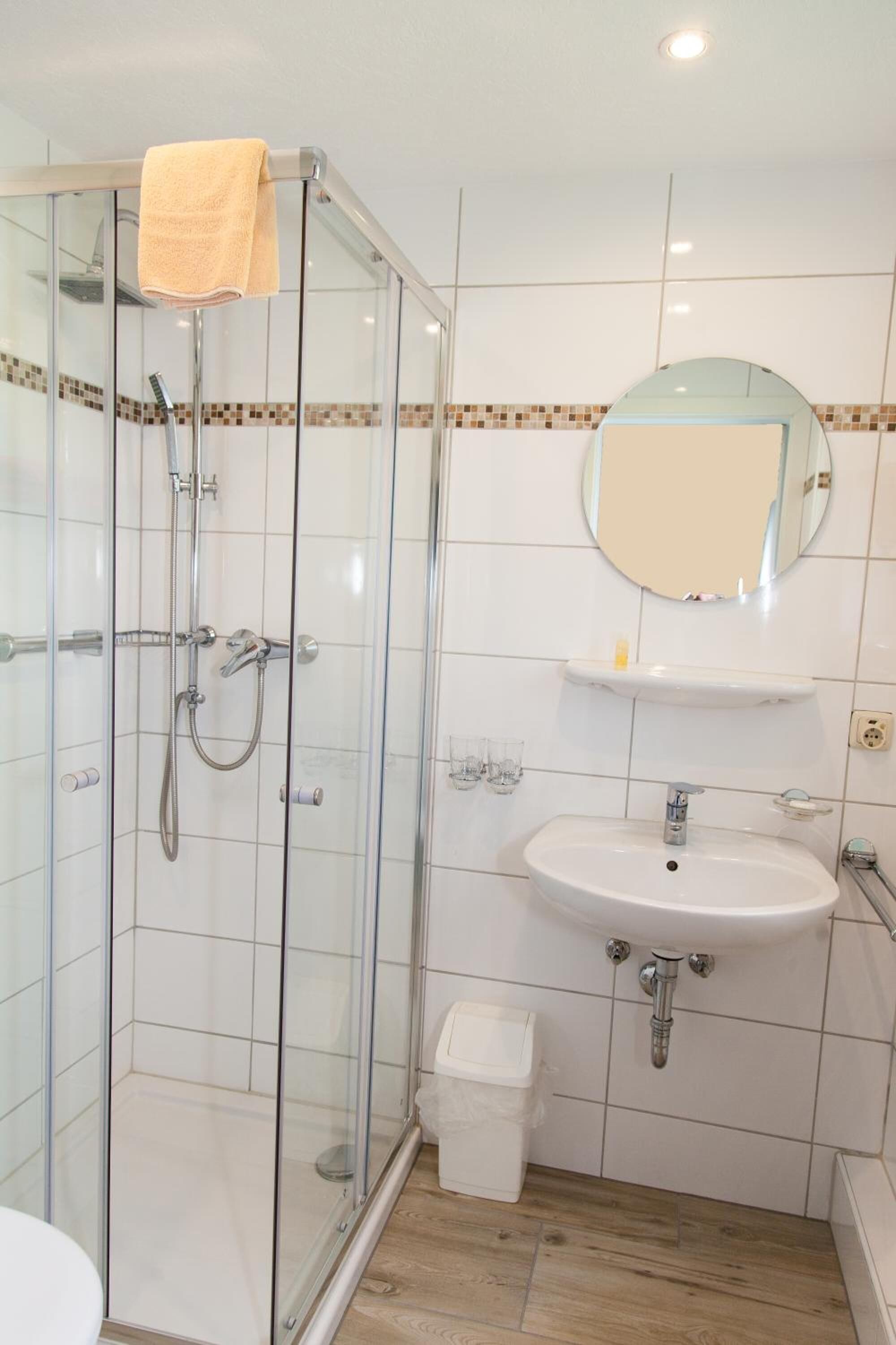 Un baño con ducha envuelta en vidrio, un lavamanos con espejo y un pequeño cubo de basura.