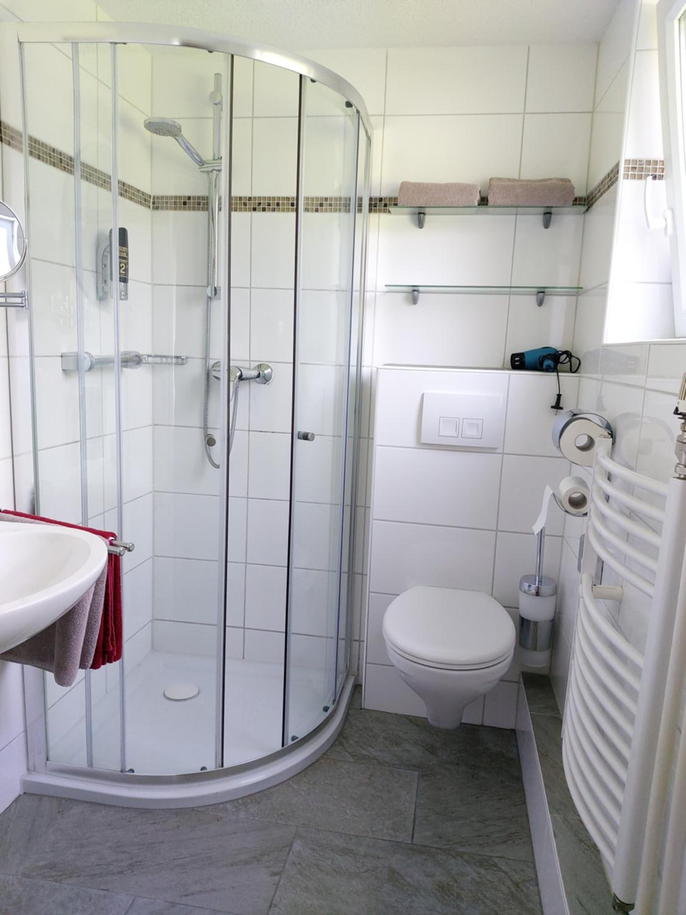 Et badeværelse med en glasafskærmet brusekabine, en hvid vask med et rødt håndklæde, et toilet og en håndklædestang.