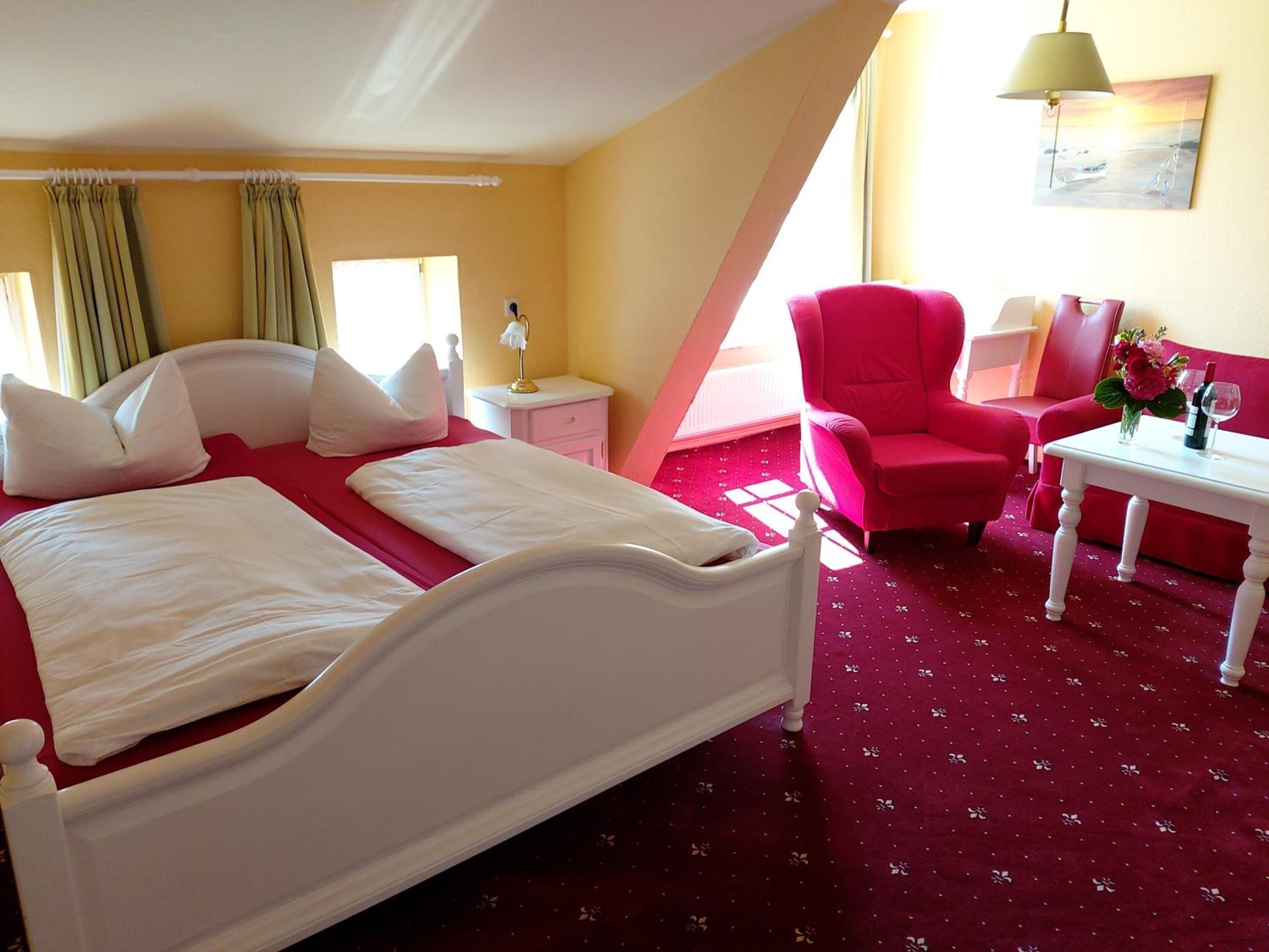 Une chambre d’hôtel avec un lit, un fauteuil rouge, une petite table sur laquelle est posé un vase de fleurs, et un tableau accroché au mur.