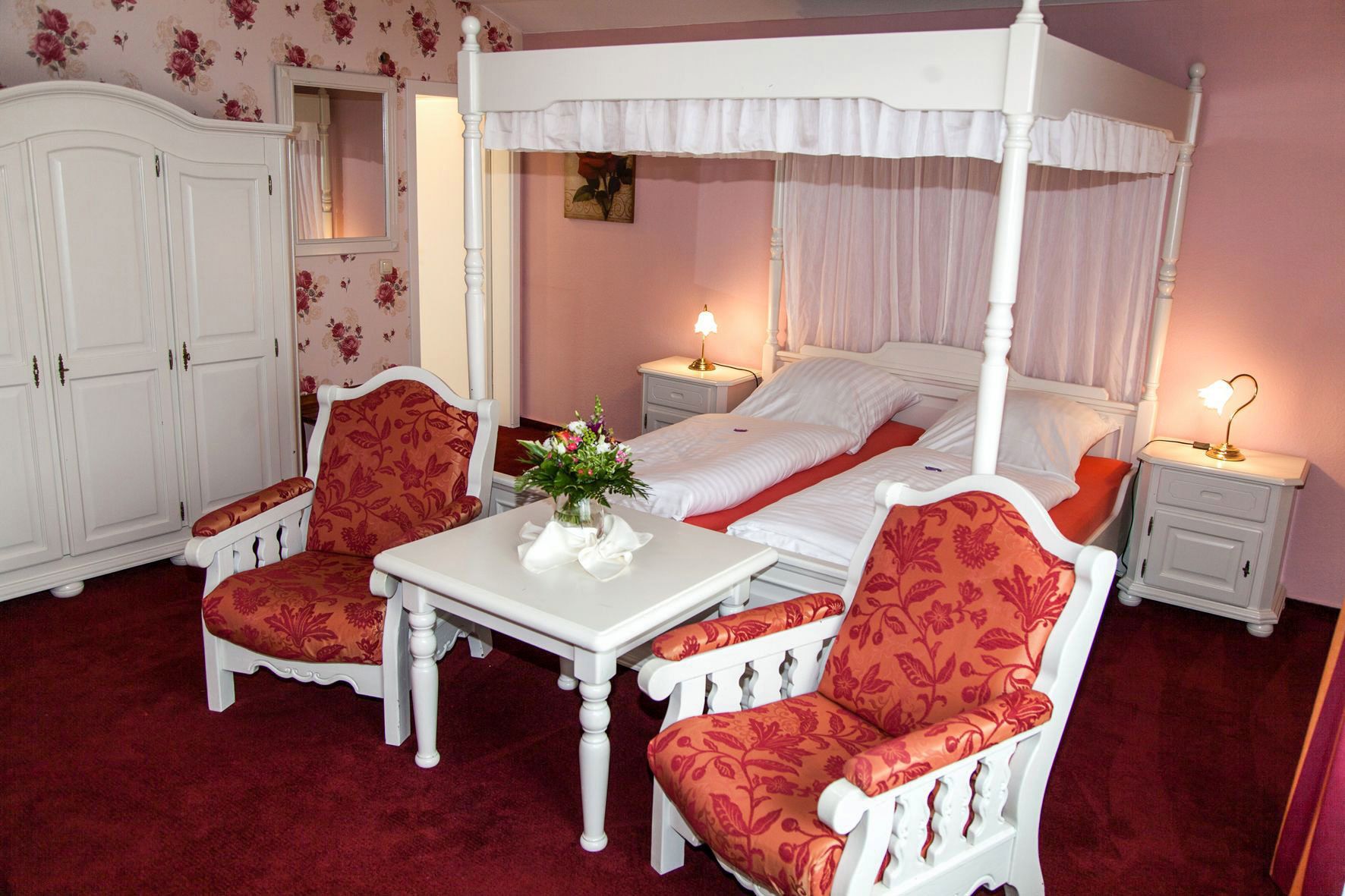 Un lit à baldaquin à quatre poteaux avec une literie blanche, deux tables de chevet avec des lampes, une table blanche et deux fauteuils rembourrés à motifs floraux.
