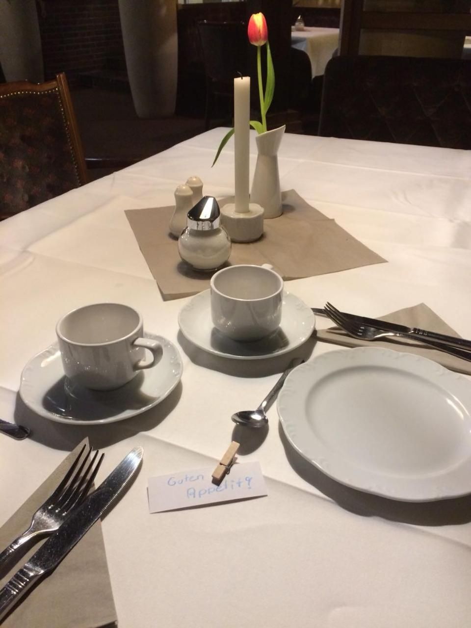 A dining table set with white plates, cups, and cutlery, a candle, and a tulip in a vase.