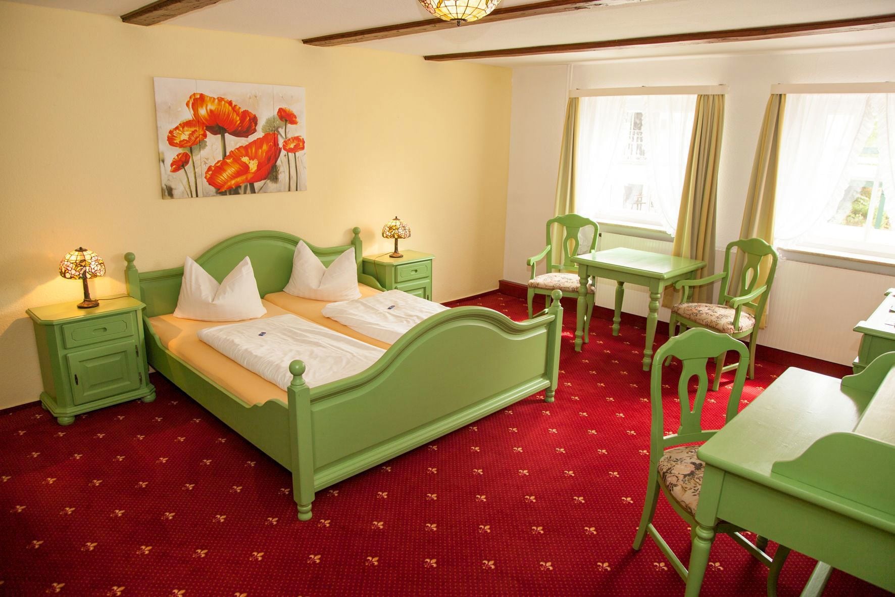 Une pièce avec un lit vert, deux tables de chevet, un bureau vert et une chaise. Un tableau représentant des fleurs est accroché au mur.