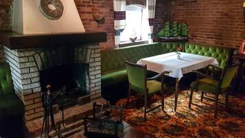 A cozy room with a brick fireplace, a green upholstered bench, a table with chairs, and a patterned carpet.