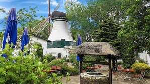 A traditional white building with a conical roof, labeled as a hotel and restaurant, surrounded by lush greenery and outdoor seating. - Hotel Der Muehlenhof (Westerdeichstrich)