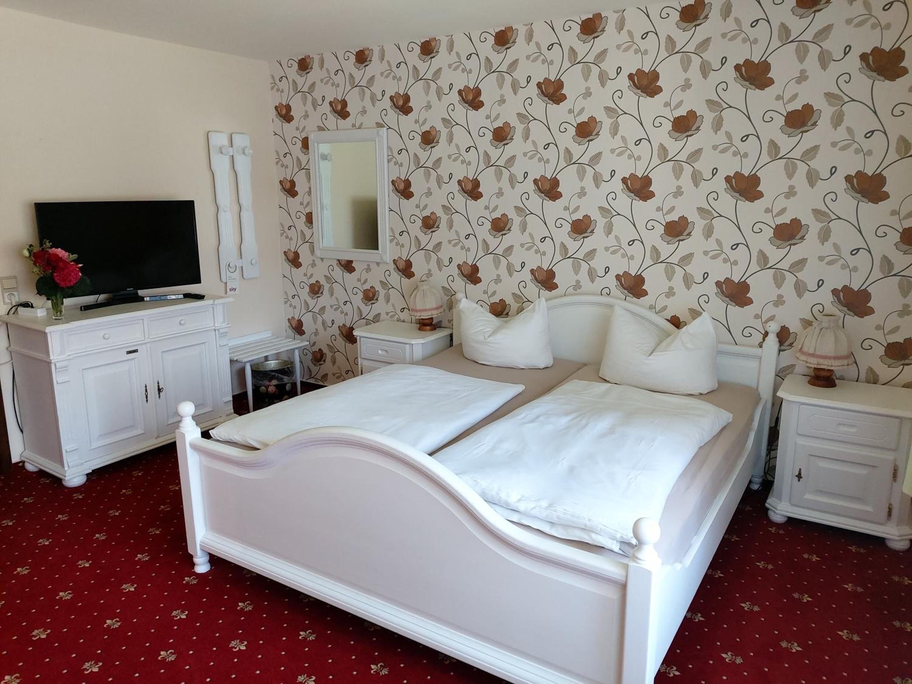 A double bed with white bedding, a TV on a white cabinet, a mirror above the bed, and a floral wallpaper.