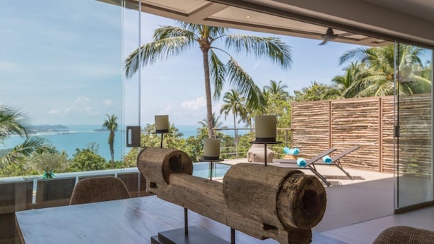A modern dining area with a large wooden sculpture centerpiece, a glass table, and a view of the ocean and palm trees. - Villa Nuéa at Comoon Boutique Villas (Koh Samui)