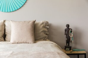 A bed with beige bedding and pillows, a small table with a statue and a woven mat beside it.