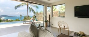 A modern living room with a large window overlooking a beach and palm trees.
