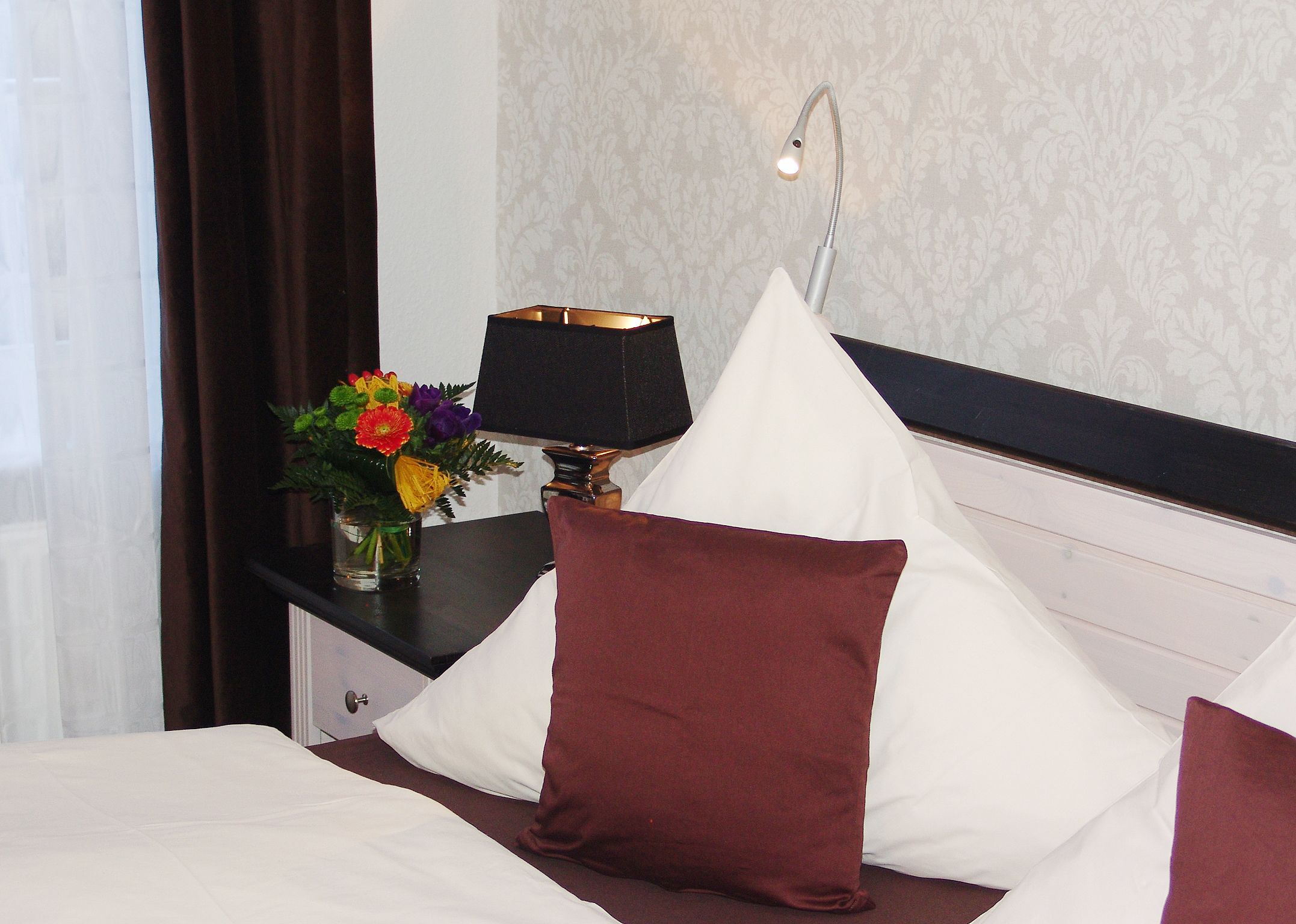 A neatly made bed with white and maroon pillows, a small table with a lamp and a vase of flowers, and a wall with a subtle pattern.