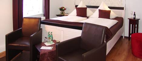 A hotel room with a bed, two armchairs, a small table with bottles, and a nightstand with a lamp.