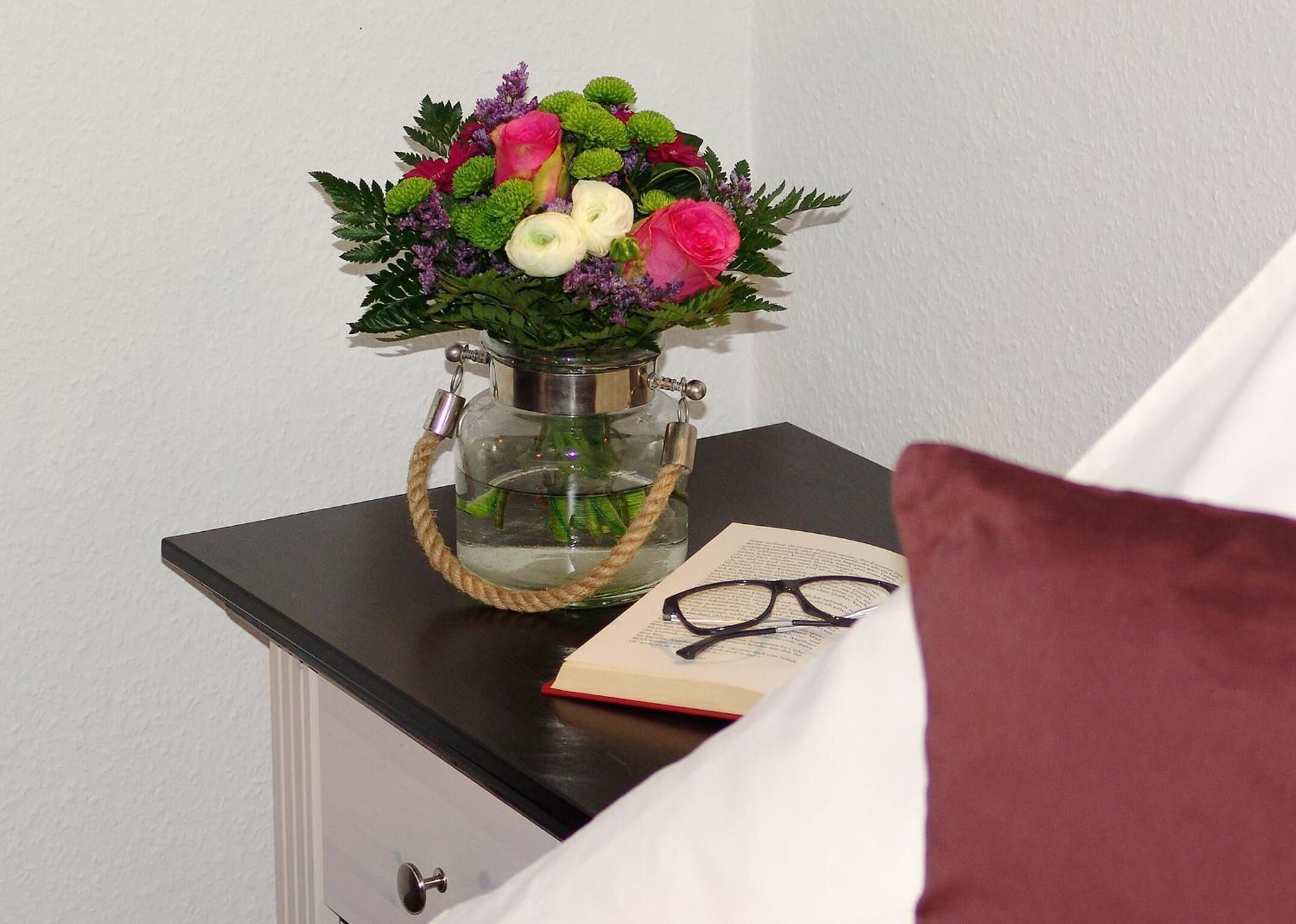 A bedside table with a glass vase containing a bouquet of flowers, a book, and a pair of glasses.