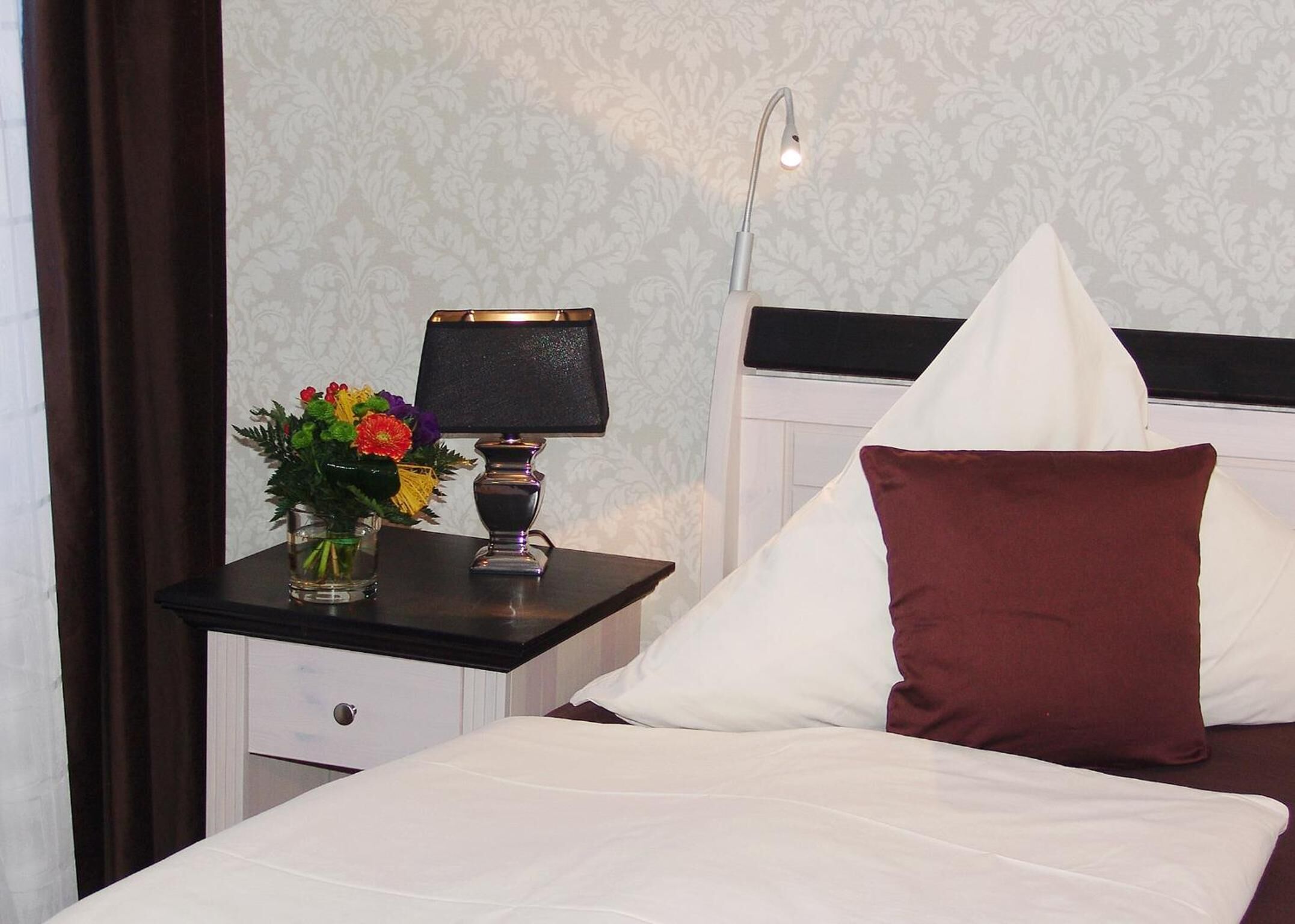 A neatly made bed with white and maroon pillows, a bedside table with a lamp and a vase of flowers, and a wall with a subtle floral pattern.