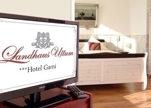 A hotel room with a television displaying 'Landhaus Utten Hotel Garni' and a bed with white bedding and a brown pillow. - Landhaus Uttum (Krummhoern)