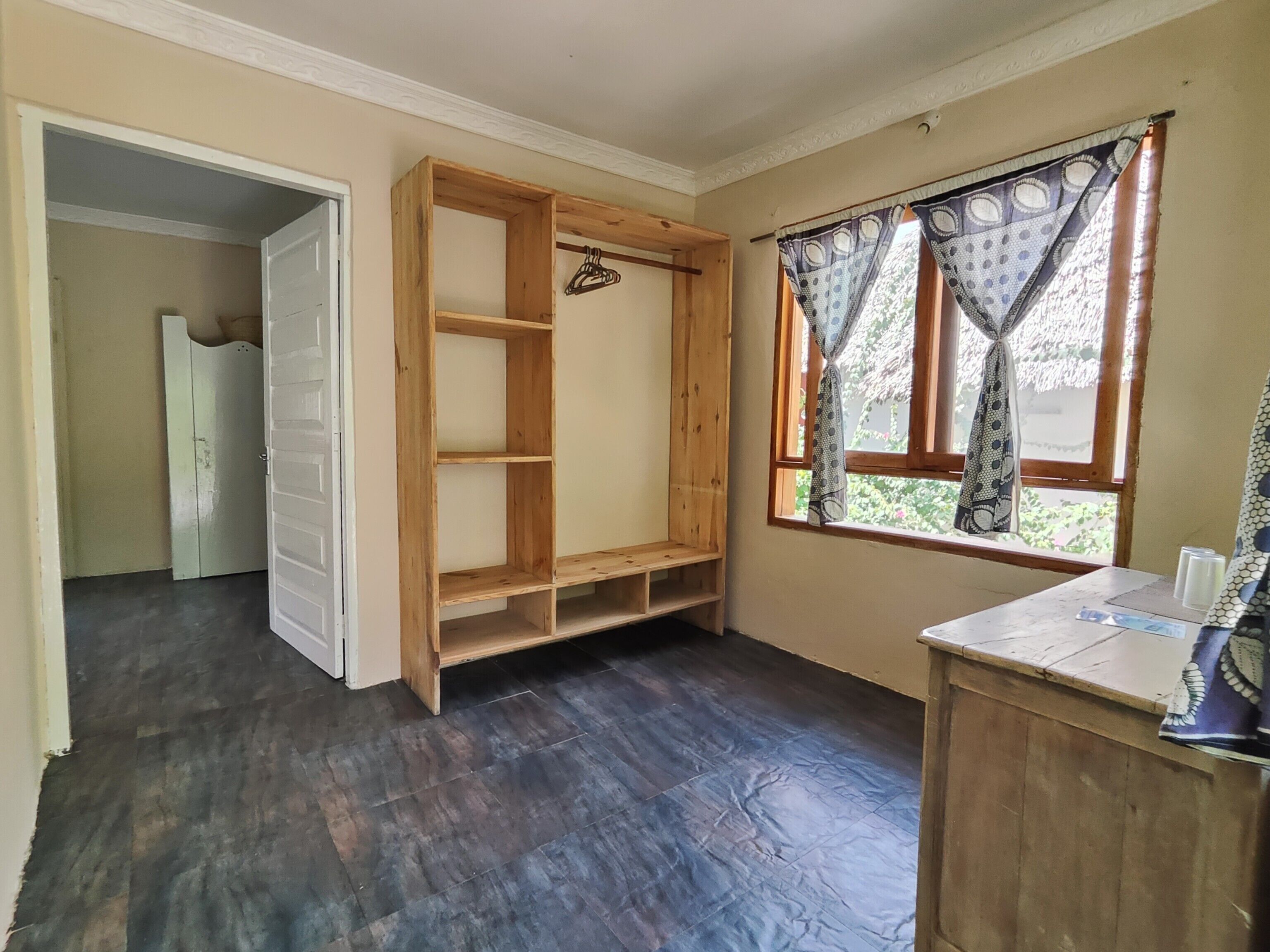 Ein Zimmer mit Holzschrank, kleinem Tisch mit Vase und Fenster mit gemusterten Vorhängen.