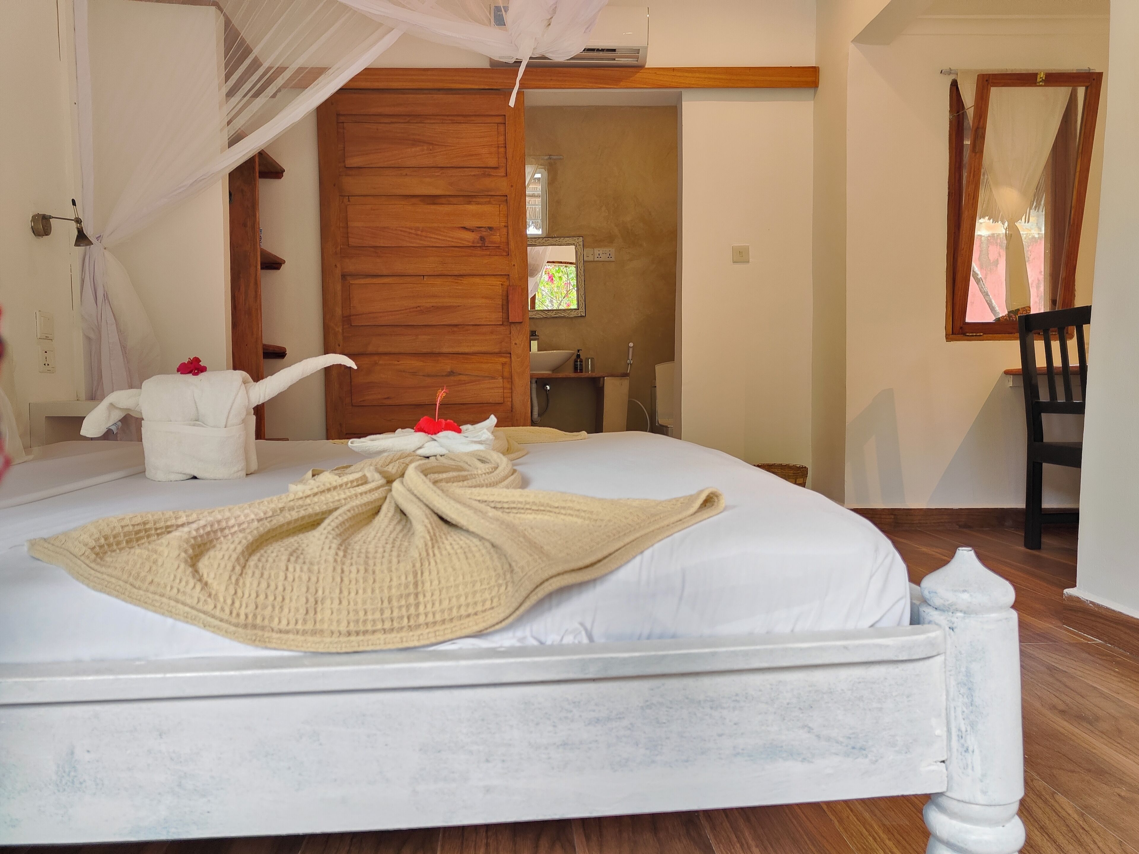 Una cama bien hecha con una colcha blanca y un edredón color beige, una puerta de madera, una mesita con artículos de tocador y una silla.
