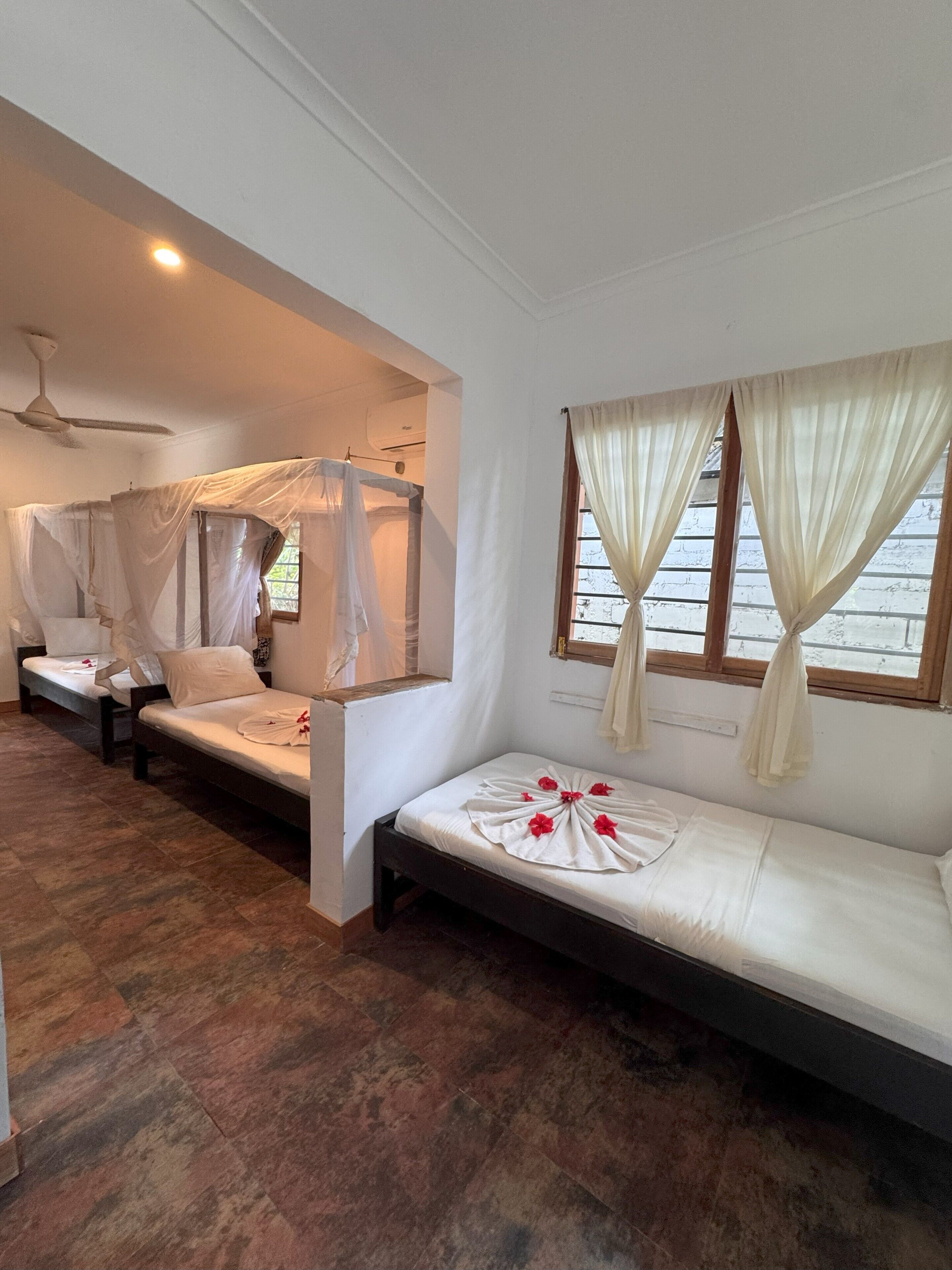 Una habitación con dos camas, una con dosel y otra sin él. Tiene un ventilador de techo, una ventana con cortinas y suelo de madera.