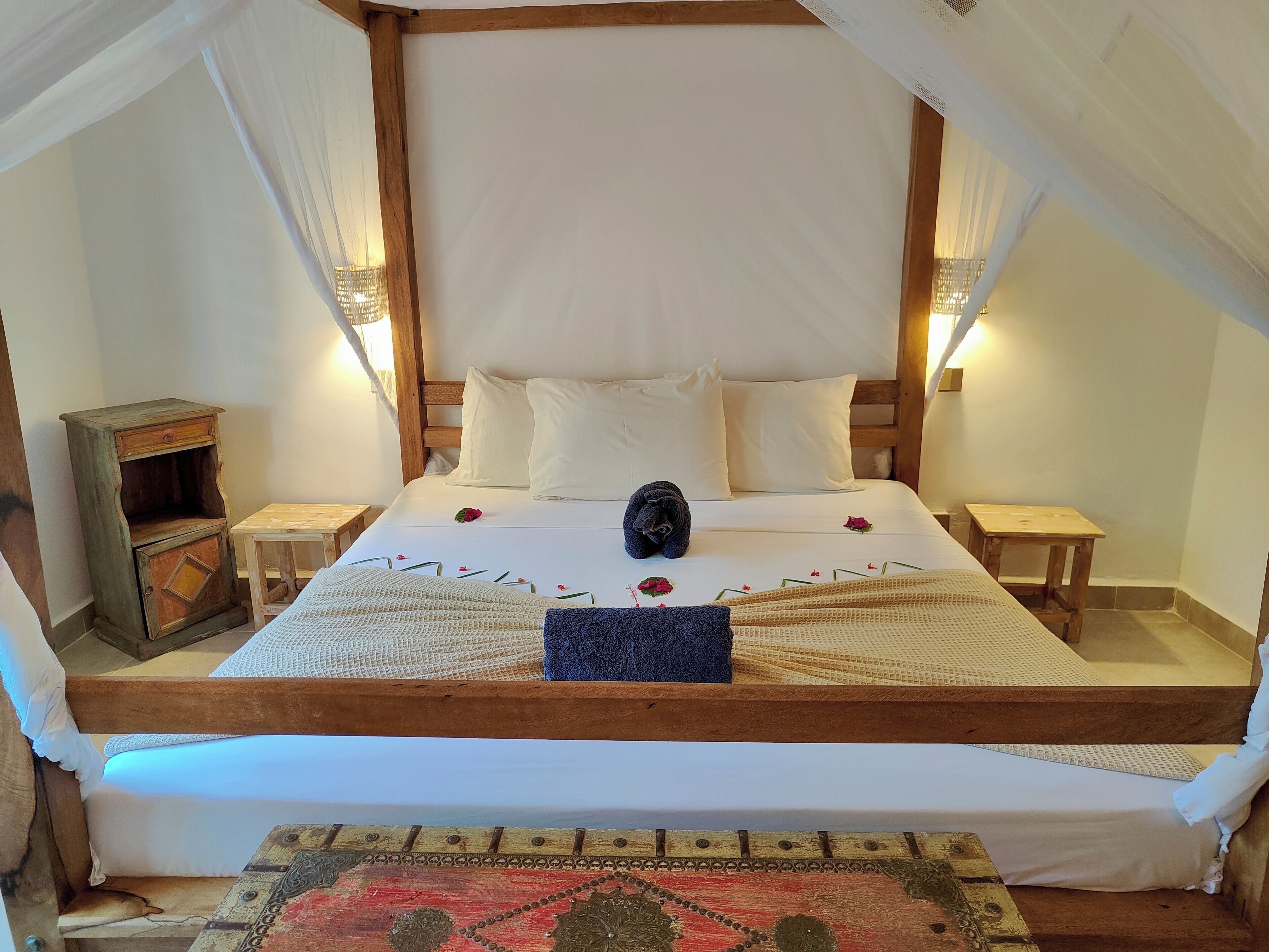 A bed with a canopy, white bedding, and a decorative pillow. There are two bedside tables, one with a lamp and the other with a small wooden box.
