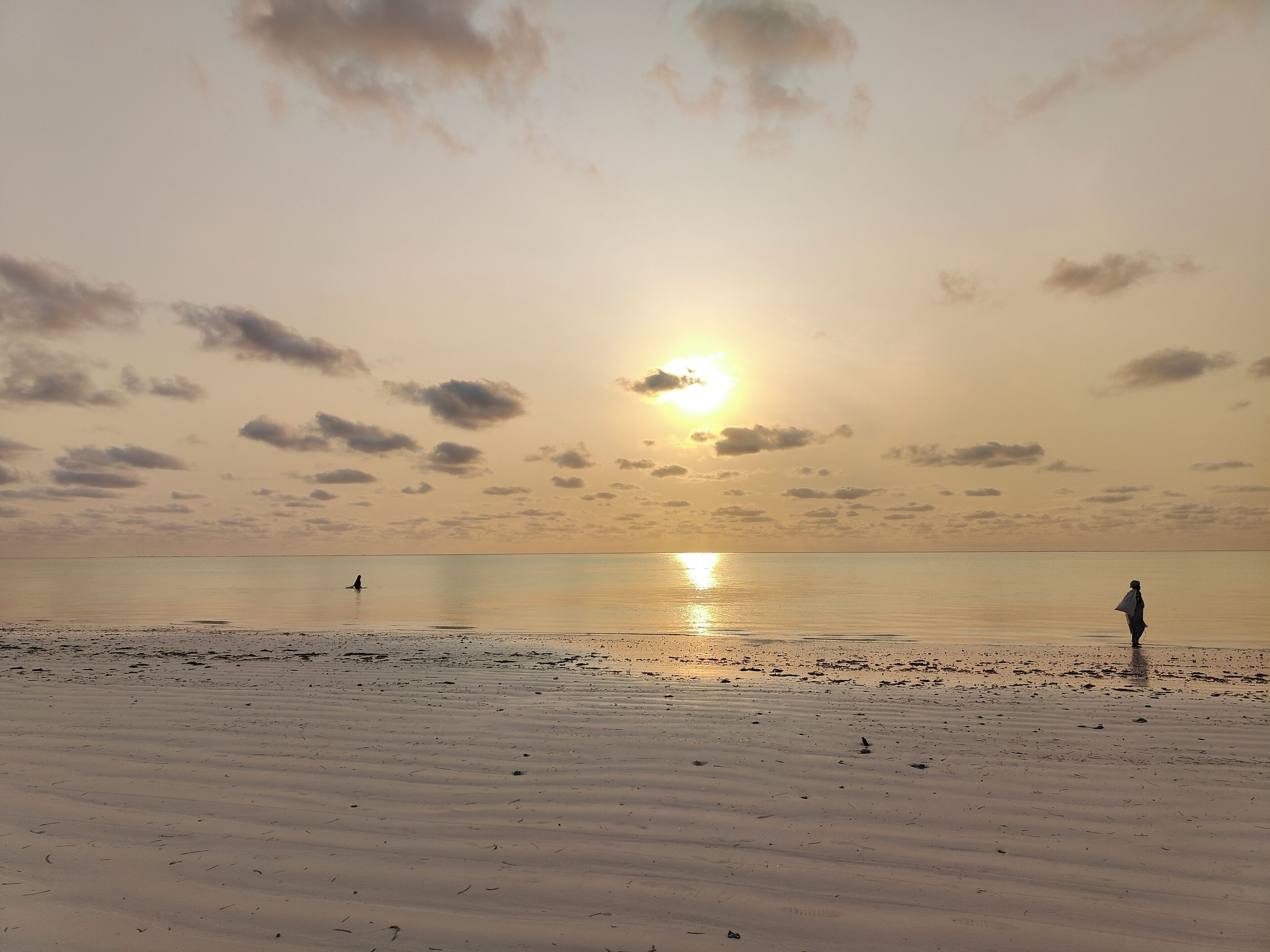 Una playa con una persona parada a lo lejos, el sol poniéndose en el horizonte y agua tranquila.