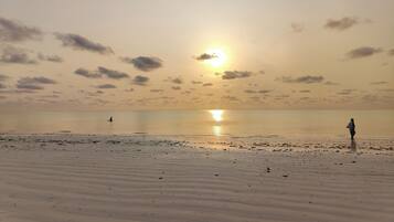 Una playa con una persona parada a lo lejos, el sol poniéndose en el horizonte y agua tranquila.