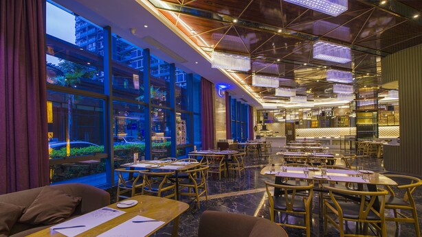 Un restaurante con varias mesas y sillas, amplias ventanas con vistas al exterior y un diseño de techo moderno con lámparas colgantes.