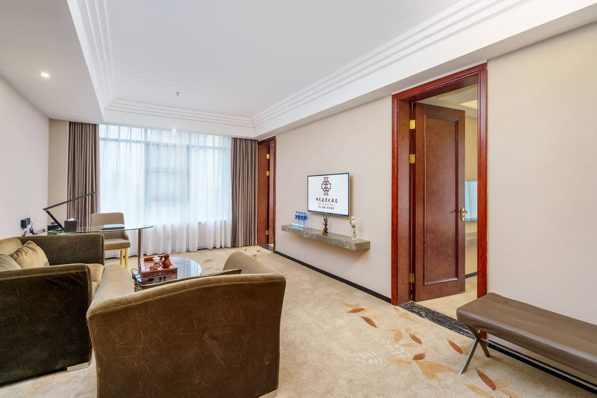 Ein modernes Hotelzimmer mit einer Couch, einem Sessel und einem kleinen Tisch mit Tablett. Eine Holztür und ein gerahmtes Bild sind an der Wand zu sehen.