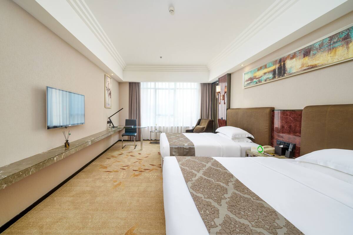 Ein Hotelzimmer mit zwei Betten, einem Schreibtisch, einem Stuhl, einem Fernseher und einem großen Fenster.