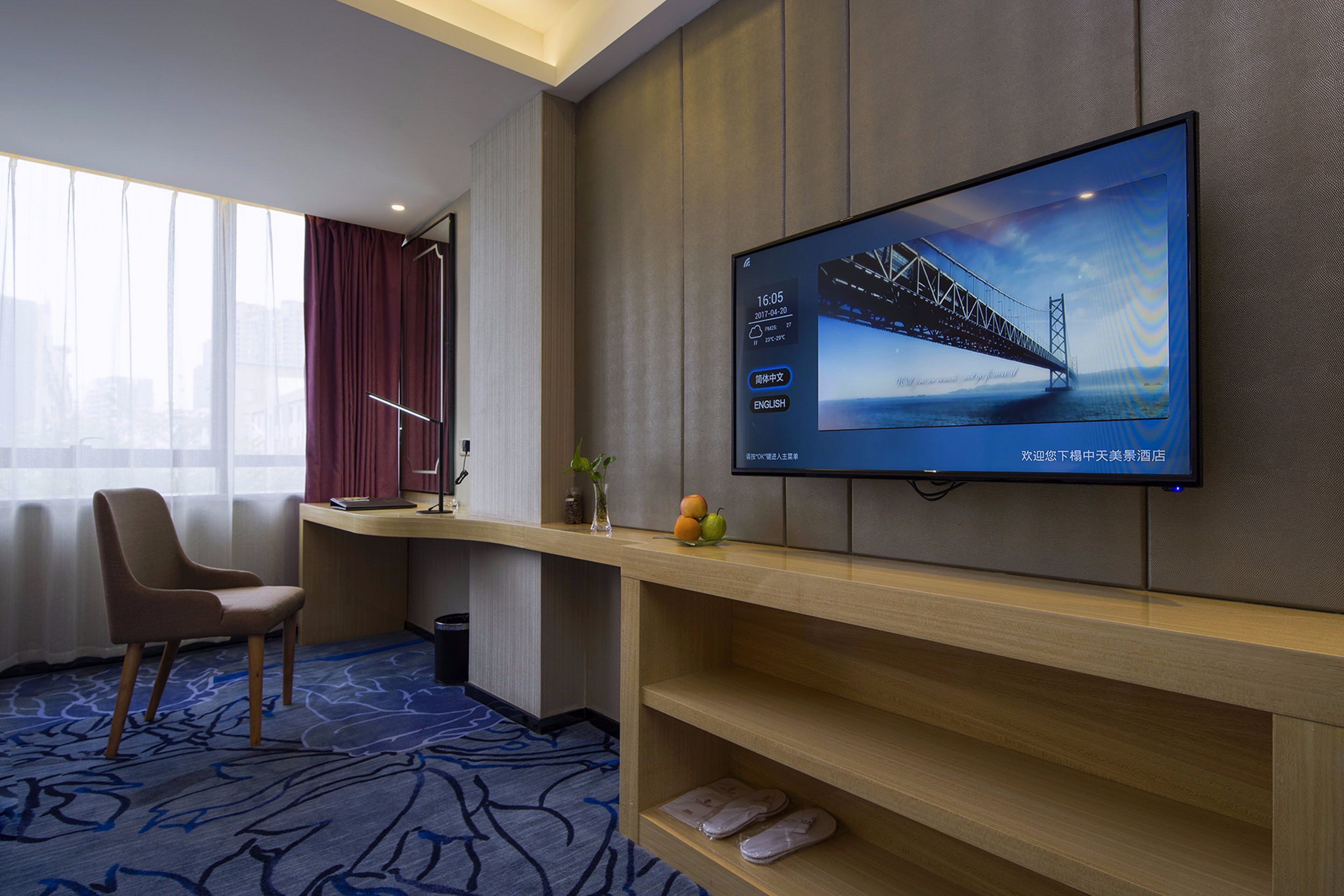 A hotel room with a large TV displaying a bridge, a desk with a vase and fruit, a chair, and a carpeted floor.