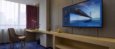 A hotel room with a large TV displaying a bridge, a desk with a vase and fruit, a chair, and a carpeted floor.