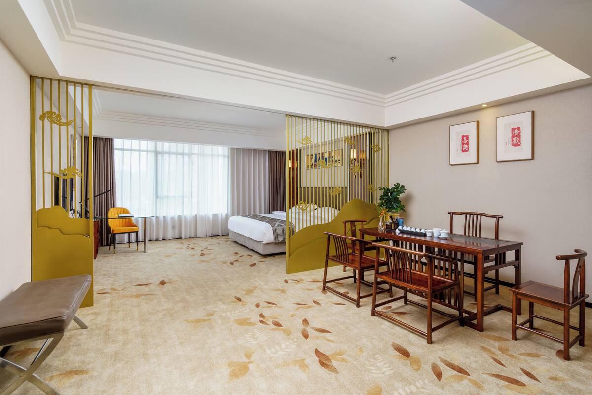 Ein Hotelzimmer mit einem Bett, einem Esstisch mit Stühlen, einem kleinen Tisch mit Pflanze und einer Bank.