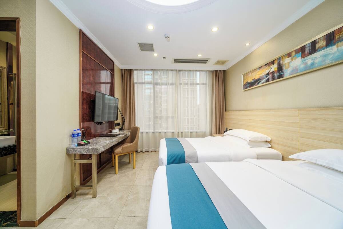 Ein Hotelzimmer mit zwei Betten, einem Schreibtisch mit Fernseher, einem Sessel und einem großen Fenster mit Vorhängen.