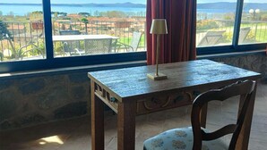 A room with a table and chair, a lamp, and a view of a body of water and landscape through large windows. - Le vigne (Bolsena)