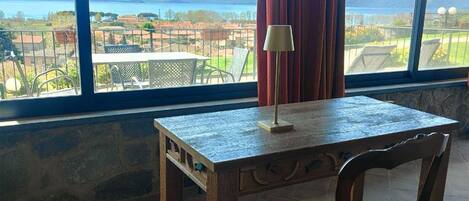 A room with a table and chair, a lamp, and a view of a body of water and landscape through large windows.