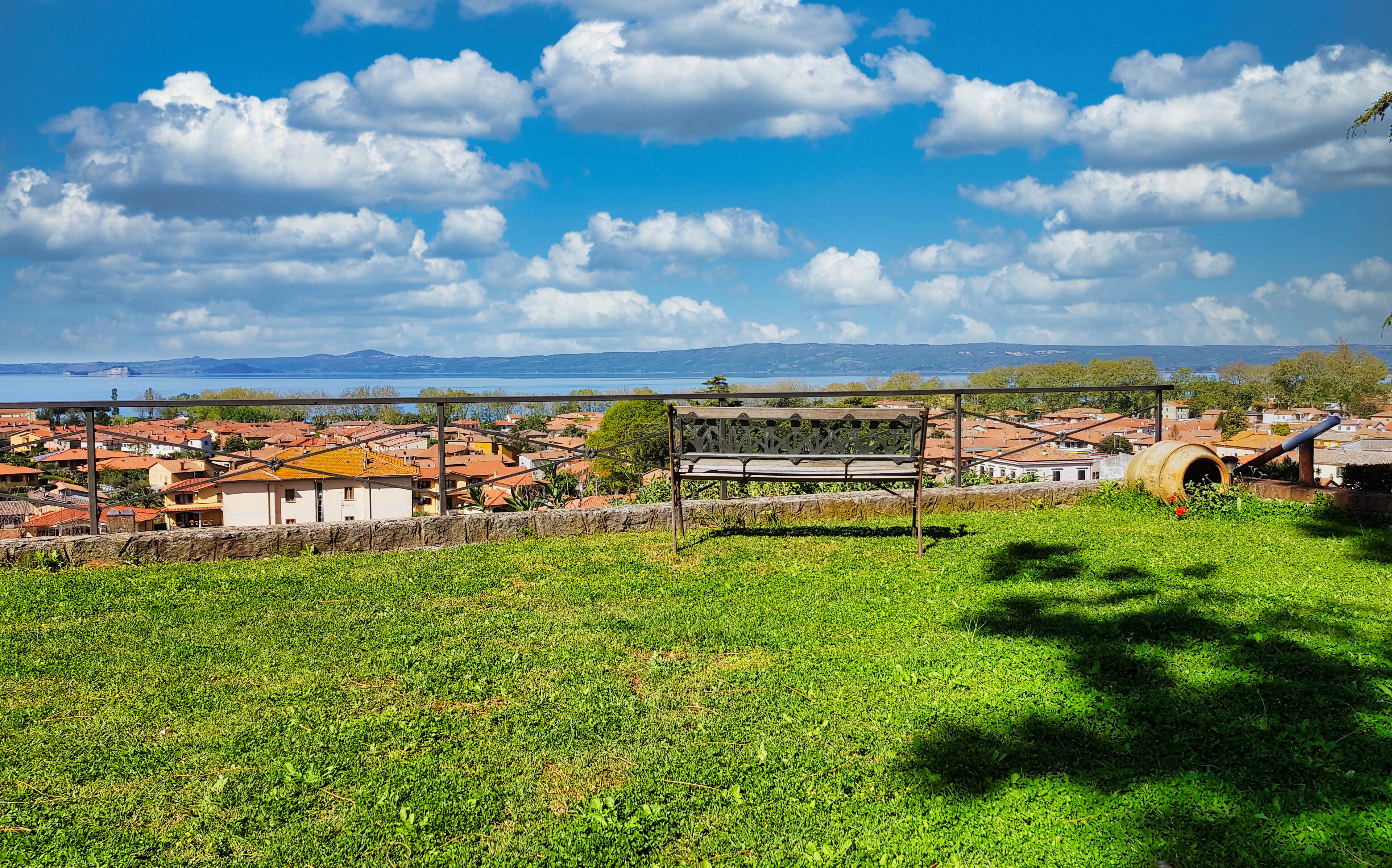 Zona verde con un banco y vista a un pueblo con tejados rojos y una masa de agua distante.