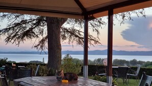 A covered patio with a table and chairs, overlooking a body of water and distant mountains. - Le vigne (Bolsena)