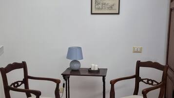 A room with two wooden chairs, a small table with a blue lamp, and two framed pictures on the wall.